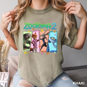 May include: Khaki-colored t-shirt featuring the text "ZOOTOPIA 2" above a graphic of characters from the movie. The graphic includes a sloth, fox, rabbit, and snake, each in a colorful frame. The shirt is a comfortable, casual style.