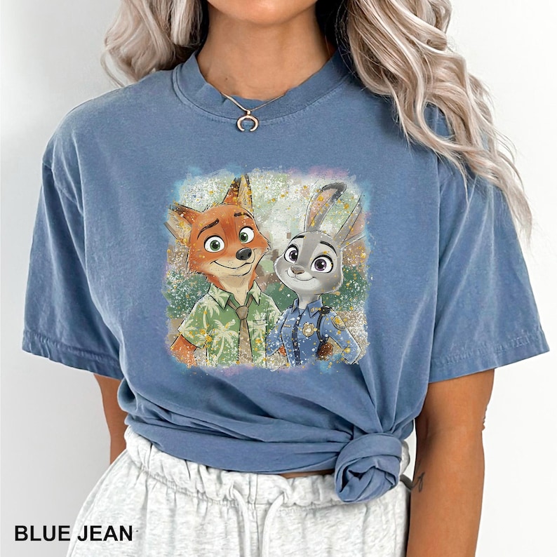 May include: Blue jean colored t-shirt featuring a colorful graphic of Nick Wilde and Judy Hopps from Zootopia. The graphic shows the characters smiling. The text "BLUE JEAN" is at the bottom of the shirt.