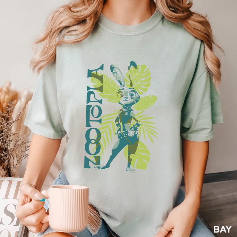 May include: A light green t-shirt featuring a graphic of Judy Hopps from Zootopia, with the word "Zootopia" in teal. The design includes a cartoon image of Judy Hopps and tropical leaves in shades of green.