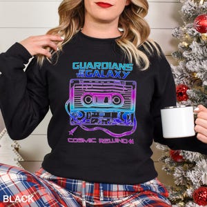 May include: Black sweatshirt featuring a colorful graphic of a cassette tape with the text "GUARDIANS OF THE GALAXY" and "COSMIC REWIND". The design uses blue, purple, and pink hues. The word "BLACK" is printed at the bottom left.