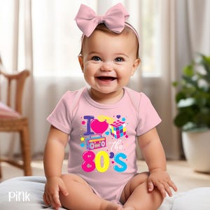 May include: A light pink short-sleeved bodysuit featuring a colorful graphic print that includes a heart, a Rubik's cube, a boombox, and the text "I love the 80's" in bright blue, yellow, and pink. A matching pink bow headband is worn on the head.