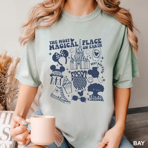 Puede incluir: Una camiseta verde claro con un gr&aacute;fico azul marino que presenta iconos de Disney y texto. El texto incluye "The Most Magical Place on Earth", "EPCOT" y "Animal Kingdom". La camiseta tiene cuello redondo y mangas cortas.