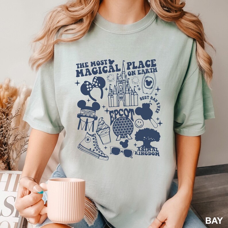 Puede incluir: Una camiseta verde claro con un gr&aacute;fico azul marino que presenta iconos de Disney y texto. El texto incluye "The Most Magical Place on Earth", "EPCOT" y "Animal Kingdom". La camiseta tiene cuello redondo y mangas cortas.