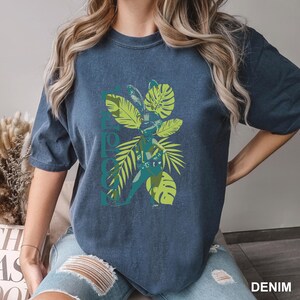 May include: A denim blue t-shirt featuring a graphic of a rabbit-like figure with green and blue tropical leaves. The word "HAPPY" is printed vertically on the left side of the shirt. The word "DENIM" is printed on the bottom right.