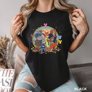 K&ouml;nnte beinhalten: Schwarzes T-Shirt mit Rundhalsausschnitt und farbenfrohem Aufdruck von Charakteren aus dem Animationsfilm Zootopia. Das Design zeigt Judy Hopps, Nick Wilde und andere Charaktere vor einem Regenbogen- und Blumenhintergrund. Das Wort "BLACK" ist unten aufgedruckt.