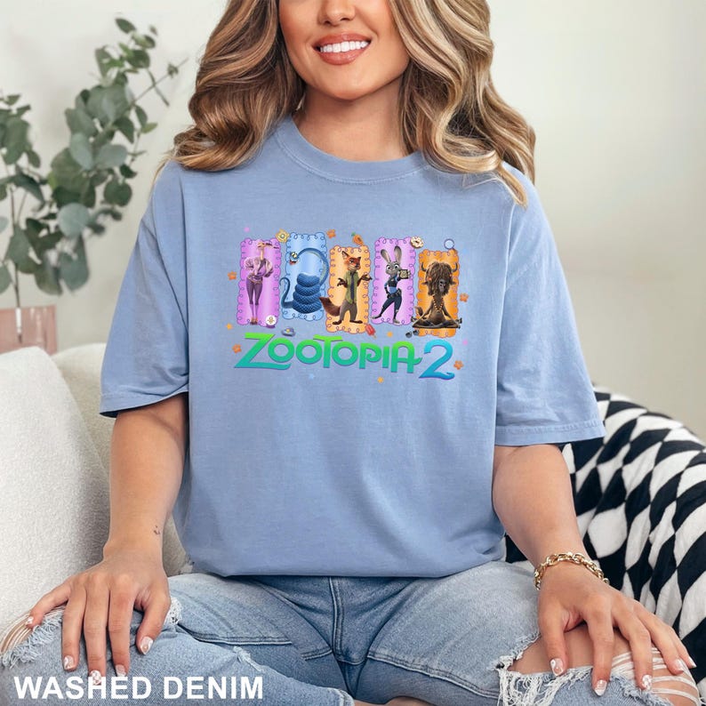 K&ouml;nnte beinhalten: Hellblaues T-Shirt mit einem farbenfrohen Grafikdesign von "Zootopia 2". Das Design zeigt Charaktere in dekorativen Rahmen, mit dem Filmtitel in Gr&uuml;n und Pink. Das T-Shirt besteht aus einem weichen Material.