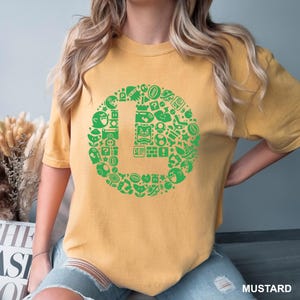 May include: Mustard-colored t-shirt featuring a large green "L" design composed of various video game icons. The design includes characters and symbols from a popular video game franchise. The shirt is a casual wear item.