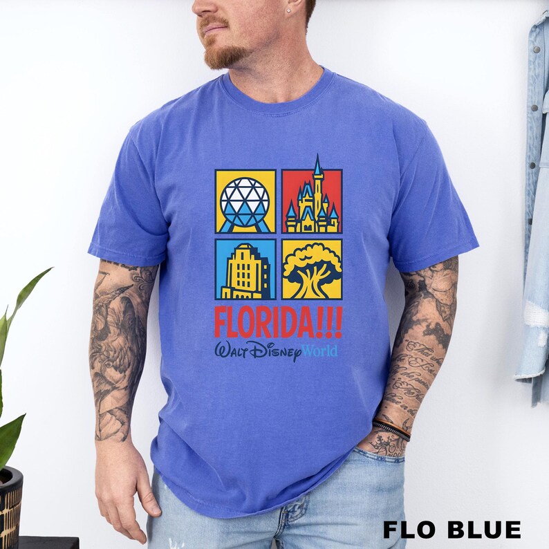 Może przedstawiać: Niebieska koszulka z kolorową grafiką przedstawiającą cztery kwadraty z kultowymi zabytkami Florydy. Słowo "FLORIDA!!!" jest wydrukowane na czerwono, z napisem "Walt Disney World" poniżej. Koszulka jest oznaczona jako "FLO BLUE" na dole.