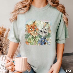 May include: A light green t-shirt featuring a colorful illustration of Nick Wilde the fox and Judy Hopps the rabbit from the animated movie Zootopia. Nick is wearing a green shirt and Judy is in a police uniform.