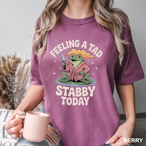 May include: Berry-colored t-shirt featuring a cartoon frog in a pink robe holding a knife. The frog sits on a lily pad with the text "Feeling a tad stabby today". The shirt has short sleeves and a crew neck.