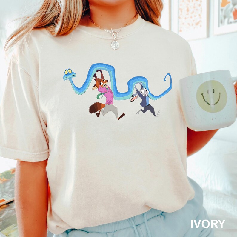 Op de afbeelding: Ivoor t-shirt met een cartoonafbeelding van een vos en een konijn die rennen met een blauwe slang. De vos draagt een roze shirt, het konijn een blauw pak. Een witte mok met een smiley wordt in de hand gehouden. Het woord "IVORY" staat onderaan.