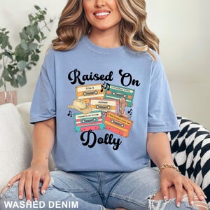 May include: A light blue t-shirt featuring the text Raised On Dolly in black script. The graphic includes four colorful cassette tapes labeled 9 to 5, Coat of Many Colors, Jolene, Islands in the Stream, and I Will Always Love You, with a cowboy hat and boot.