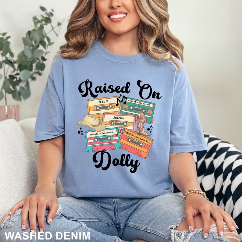 May include: A light blue t-shirt featuring the text Raised On Dolly in black script. The graphic includes four colorful cassette tapes labeled 9 to 5, Coat of Many Colors, Jolene, Islands in the Stream, and I Will Always Love You, with a cowboy hat and boot.