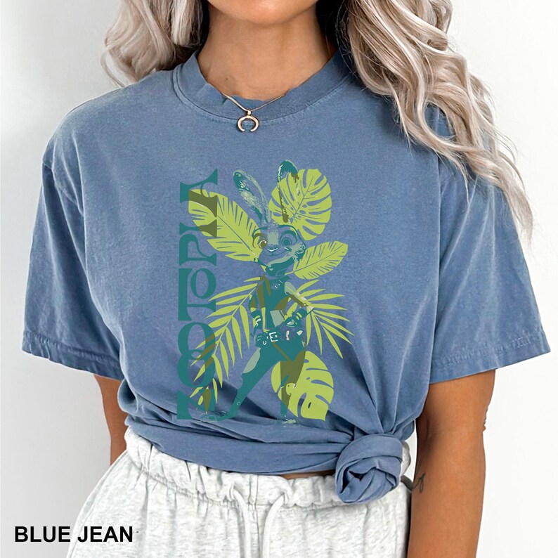 May include: Blue jean colored t-shirt featuring a cartoon image of a rabbit character from Zootopia, surrounded by green tropical leaves. The word "ZOOTOPIA" is printed vertically in green. The shirt is tied at the waist.