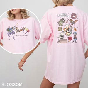 May include: Pale pink t-shirt with cartoon characters and the text "You can be anything you wish to be." The back of the shirt has a graphic of cartoon characters and the text "The Big Donut."