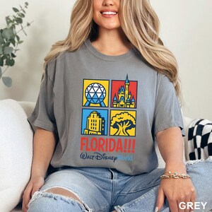 Może przedstawiać: Szara koszulka z kolorową grafiką czterech kwadrat&oacute;w z symbolami Florydy. Słowo "FLORIDA!!!" jest wydrukowane na czerwono, z napisem "Walt Disney World" poniżej. Koszulka ma wygodny kr&oacute;j.