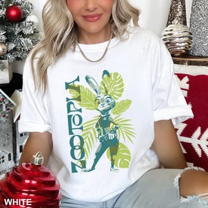 May include: White t-shirt featuring a graphic of Judy Hopps from Zootopia. The design includes the word "Zootopia" in green, along with a cartoon image of Judy Hopps and green tropical leaves. The shirt is a casual, comfortable style.