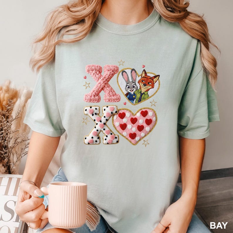 May include: A light sage green t-shirt featuring a graphic print of the characters Judy Hopps and Nick Wilde inside a heart, alongside the letters X and O in textured pink and white patterns, surrounded by small gold stars and red hearts.
