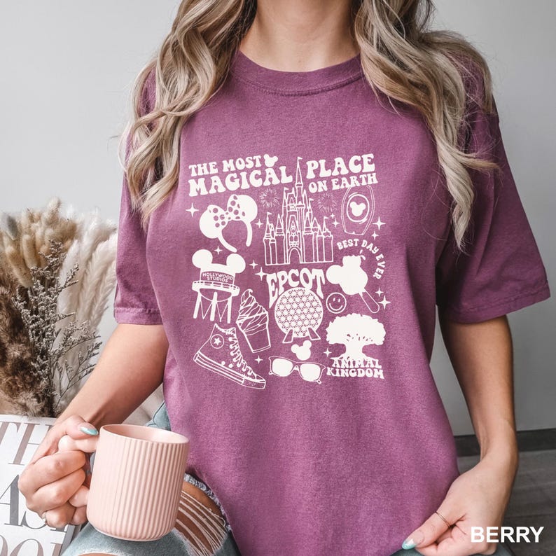 Puede incluir: Camiseta de color bayas con un dise&ntilde;o gr&aacute;fico blanco. El dise&ntilde;o incluye el texto "THE MOST MAGICAL PLACE ON EARTH" e iconos de Disney, como un castillo y orejas de Mickey Mouse. La camiseta es de cuello redondo y manga corta.