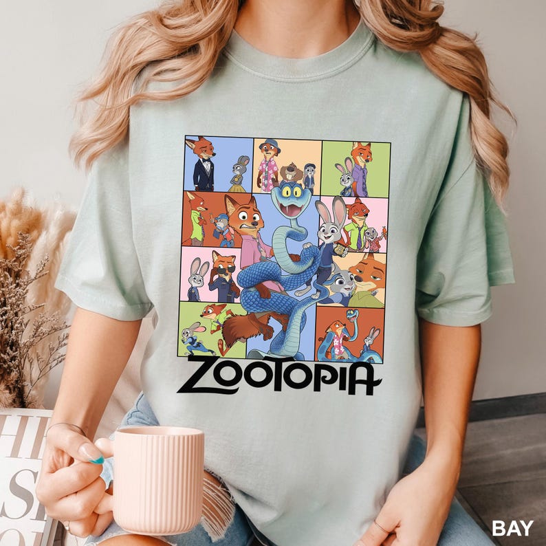 May include: A light green t-shirt featuring a colorful graphic of characters from the animated movie "Zootopia". The design includes a snake, rabbits, foxes, and other animals. The word "Zootopia" is printed in black at the bottom.