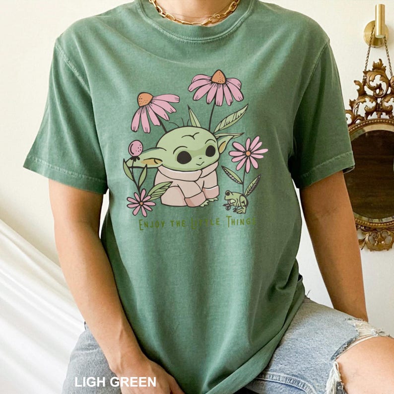 Puede incluir: Camiseta verde claro con un personaje de dibujos animados rodeado de flores rosas y una peque&ntilde;a rana. El texto "Enjoy the Little Thing" est&aacute; impreso debajo. La camiseta es de color liso.