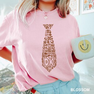 May include: A light pink t-shirt featuring a brown tie design filled with video game icons and the letters "DK". The shirt is paired with light blue shorts. A white mug with a yellow smiley face is held in the hand.