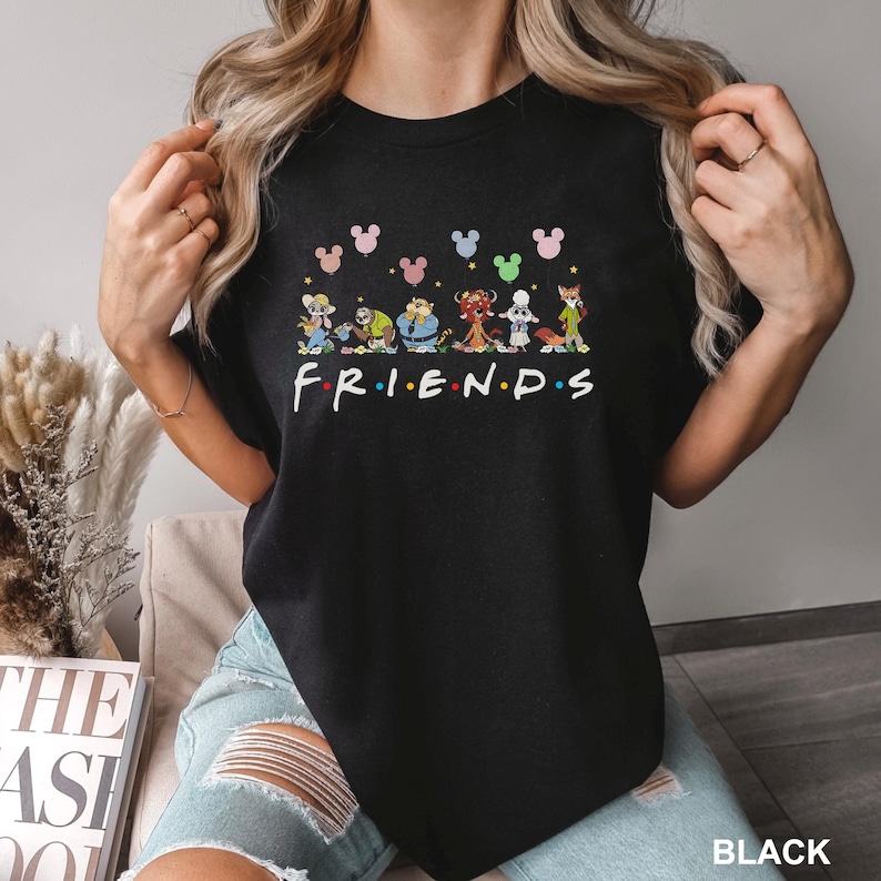 K&ouml;nnte beinhalten: Schwarzes Rundhals-T-Shirt mit einer bunten Grafik mit Zeichentrickfiguren und dem Wort "FRIENDS" in einer Regenbogen-Schriftart. &Uuml;ber den Figuren befinden sich Mickey Mouse-Kopfformen in verschiedenen Farben. Das Wort "BLACK" befindet sich unten rechts.