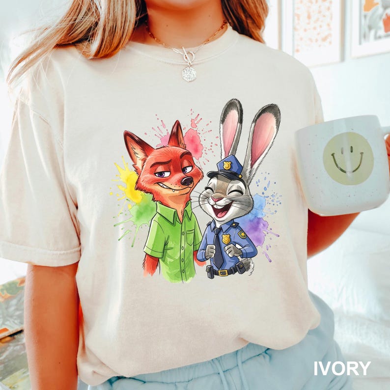 May include: Ivory-colored t-shirt featuring a colorful graphic of a fox and a rabbit, with a watercolor splash effect. The fox wears a green shirt, and the rabbit is in a police uniform. A white mug with a smiley face is held in the image.