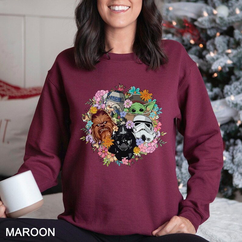 Op de afbeelding: Bordeaux sweatshirt met een bloemenontwerp met Star Wars-personages zoals Darth Vader, Chewbacca en Baby Yoda. Het ontwerp is omgeven door kleurrijke bloemen. Het woord "MAROON" staat onderaan gedrukt.