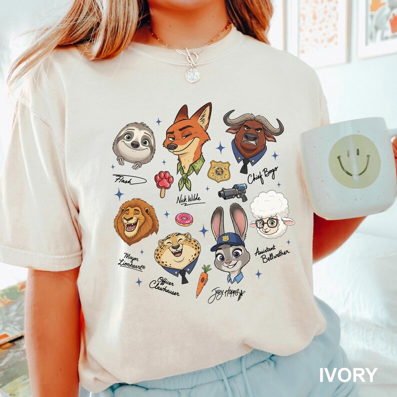 K&ouml;nnte beinhalten: Elfenbeinfarbenes T-Shirt mit Cartoon-Illustrationen von Charakteren aus einem beliebten Zeichentrickfilm. Das Design umfasst ein Faultier, einen Fuchs, einen B&uuml;ffel, einen L&ouml;wen, einen Geparden, ein Kaninchen und ein Schaf, jeweils mit ihren Namen darunter. Eine Tasse mit einem Smiley ist ebenfalls sichtbar.