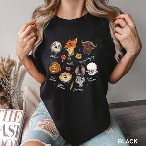 K&ouml;nnte beinhalten: Schwarzes T-Shirt mit einem farbenfrohen Aufdruck von Charakteren aus dem Animationsfilm Zoomania. Das Design enth&auml;lt Bilder von Flash, Nick Wilde, Chief Bogo, Judy Hopps und anderen. Das Wort "BLACK" ist unten rechts auf dem T-Shirt aufgedruckt.