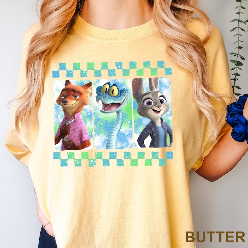 May include: Yellow t-shirt featuring a graphic of three animated characters: a fox in a pink shirt, a blue snake, and a rabbit in a blue jacket. The graphic is framed by a blue and green checkered border. The word "BUTTER" is printed at the bottom.