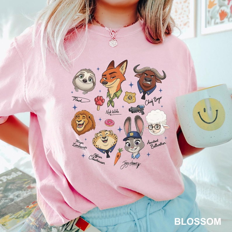 K&ouml;nnte beinhalten: Hellrosa T-Shirt mit Cartoon-Illustrationen von Charakteren aus dem Film Zootopia. Die Charaktere sind Flash, Nick Wilde, Chief Bogo, Mayor Lionheart, Officer Clawhauser, Judy Hopps und Assistant Bellwether. Eine wei&szlig;e Tasse mit gelbem Smiley ist ebenfalls zu sehen.