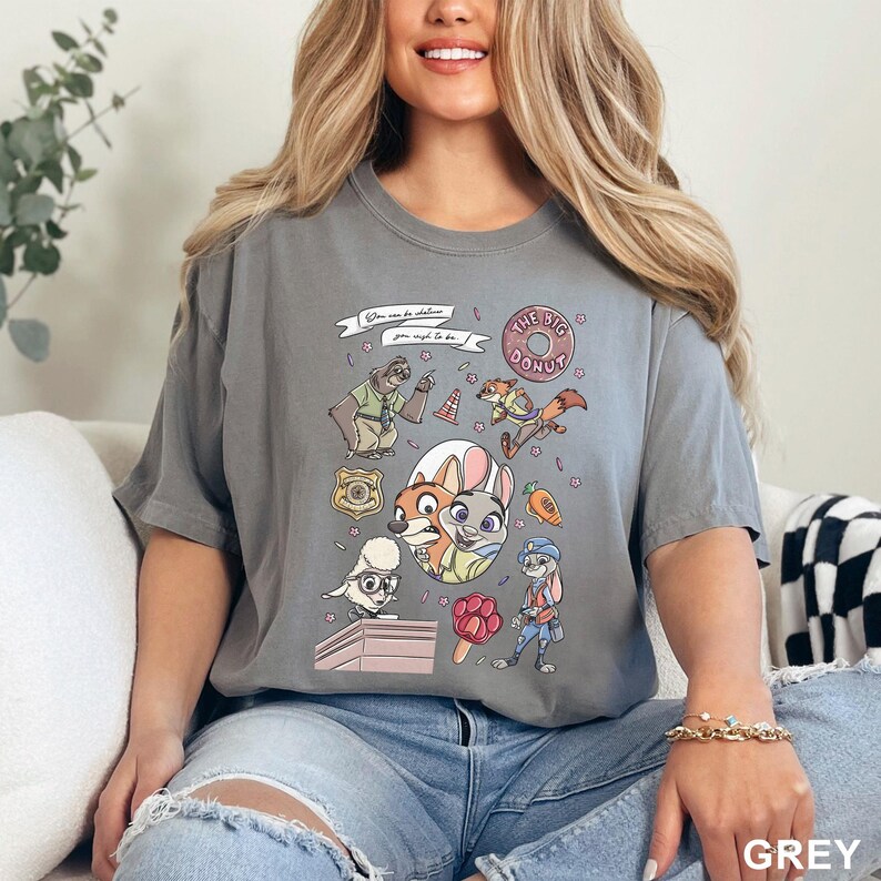Op de afbeelding: Grijze t-shirt met cartoonfiguren en de tekst "You can be whatever you wish to be." Het ontwerp bevat een donut, een politiebadge en verschillende dierenfiguren. Het woord "GREY" staat rechtsonder afgedrukt.
