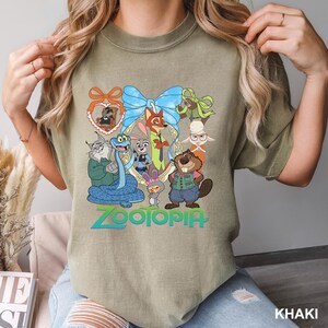 K&ouml;nnte beinhalten: Khaki-farbenes T-Shirt mit einer farbenfrohen Grafik von Charakteren aus dem Animationsfilm "Zoomania". Das Design umfasst eine blaue Schleife, einen Fuchs, ein Kaninchen, einen Biber und andere Tierfiguren. Das Wort "Zoomania" ist in Gr&uuml;n und Blau geschrieben.