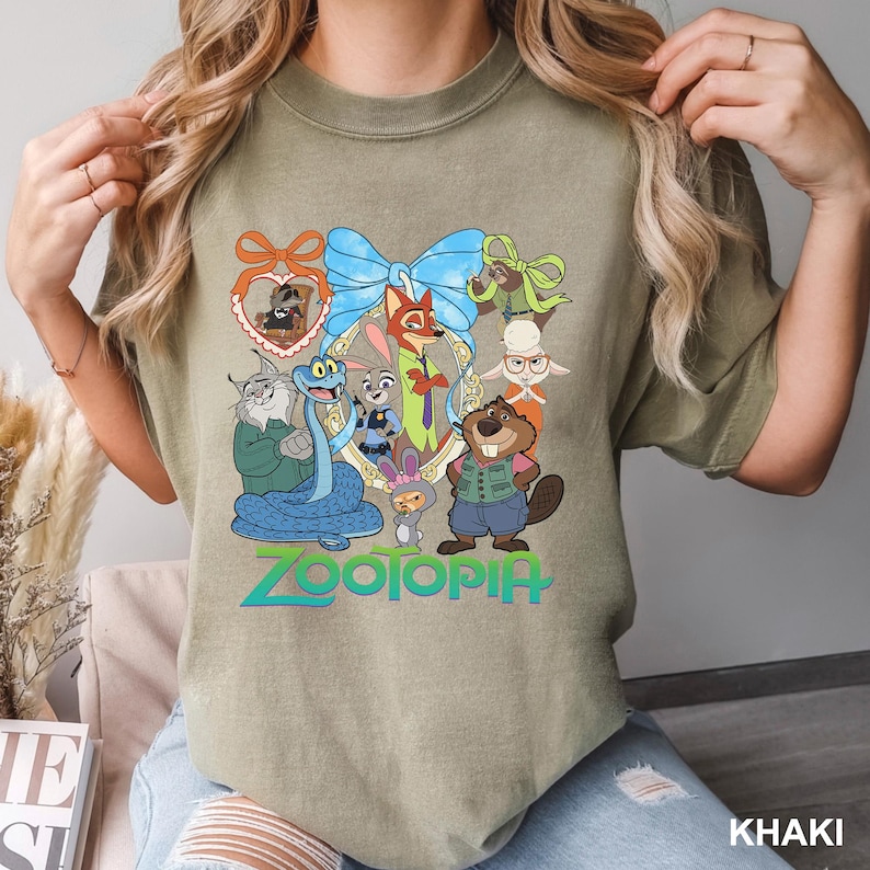 K&ouml;nnte beinhalten: Khaki-farbenes T-Shirt mit einer farbenfrohen Grafik von Charakteren aus dem Animationsfilm "Zoomania". Das Design umfasst eine blaue Schleife, einen Fuchs, ein Kaninchen, einen Biber und andere Tierfiguren. Das Wort "Zoomania" ist in Gr&uuml;n und Blau geschrieben.