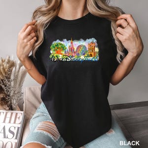 May include: A black t-shirt featuring a colorful graphic print of the Walt Disney World theme park landmarks, including the Tree of Life, Cinderella Castle, Spaceship Earth, and the Tower of Terror, with the text Walt Disney World in blue script.