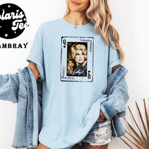 May include: A light blue crew neck t-shirt featuring a graphic of a playing card with the Queen of Hearts. The card displays a portrait of a blonde woman with the signature Dolly written below. The shirt is worn with a denim jacket and denim shorts.