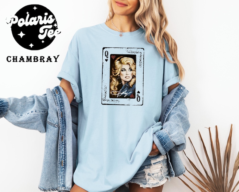 May include: A light blue crew neck t-shirt featuring a graphic of a playing card with the Queen of Hearts. The card displays a portrait of a blonde woman with the signature Dolly written below. The shirt is worn with a denim jacket and denim shorts.