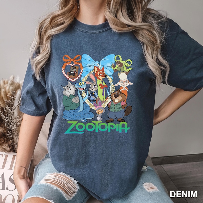 K&ouml;nnte beinhalten: Denimblaues T-Shirt mit einer farbenfrohen Grafik von Charakteren aus dem Animationsfilm "Zoomania". Das Design umfasst Judy Hopps, Nick Wilde und andere Charaktere, mit dem Wort "Zootopia" in gr&uuml;ner Schrift.
