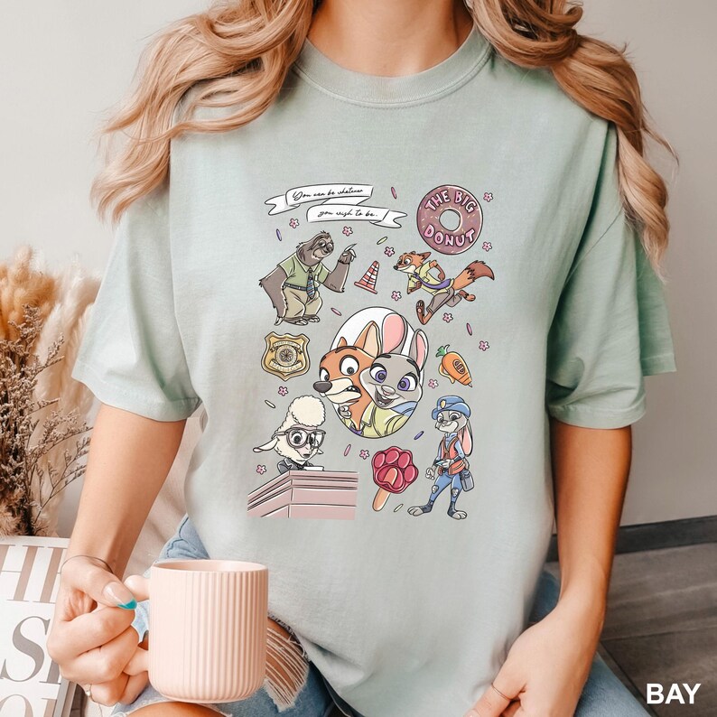 Op de afbeelding: Lichtgroen T-shirt met cartoonfiguren uit Zootopia. Het ontwerp bevat Judy Hopps, Nick Wilde en andere personages, samen met de tekst "You can be whatever you wish to be" en "The Big Donut".