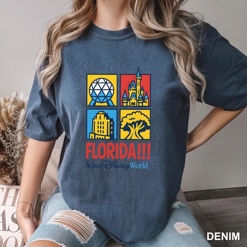 Może przedstawiać: Niebieska koszulka z dżinsu z kolorową grafiką przedstawiającą cztery kwadraty z kultowymi obrazami Florydy. Słowo "FLORIDA!!!" jest wydrukowane na czerwono, z napisem "Walt Disney World" poniżej.