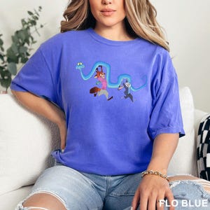 Op de afbeelding: Blauw T-shirt met een cartoon illustratie van een vos en een konijn die rennen met een blauw slangachtig ontwerp. Het shirt heeft de tekst "FLO BLUE" onderaan.