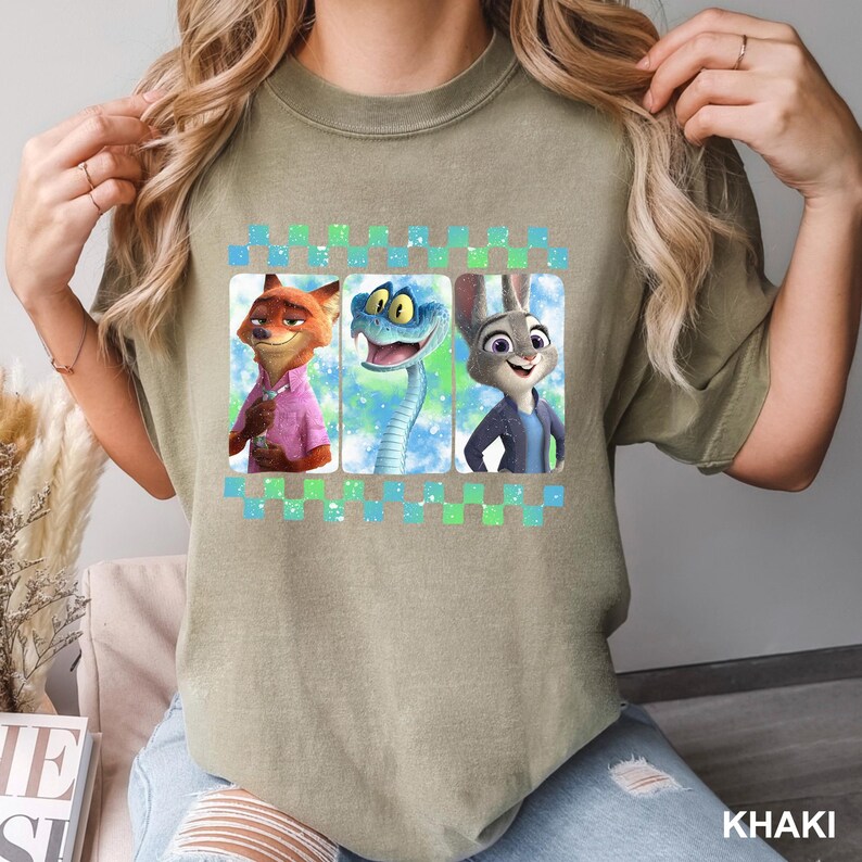 May include: Khaki-colored t-shirt featuring three cartoon characters: a fox in a pink shirt, a blue snake, and a rabbit in a blue jacket. The design is framed by a green and blue checkered border. The word "KHAKI" is printed at the bottom.