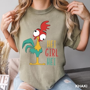 May include: Khaki-colored t-shirt featuring a cartoon rooster with red, yellow, and teal accents. The text "HEI GIRL HEI" is printed in colorful letters. The shirt is a casual wear item.
