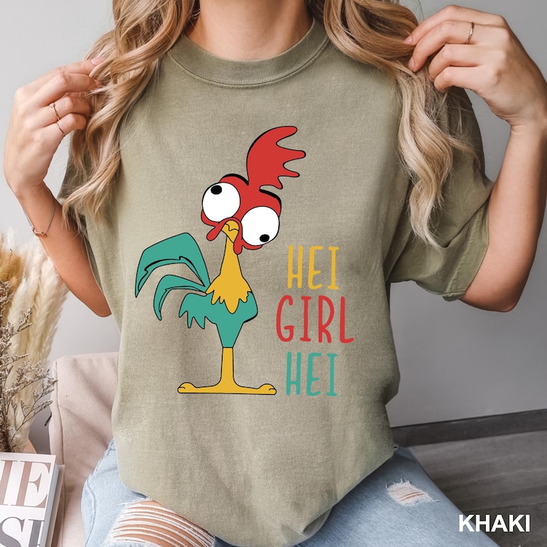 May include: Khaki-colored t-shirt featuring a cartoon rooster with red, yellow, and teal accents. The text "HEI GIRL HEI" is printed in colorful letters. The shirt is a casual wear item.