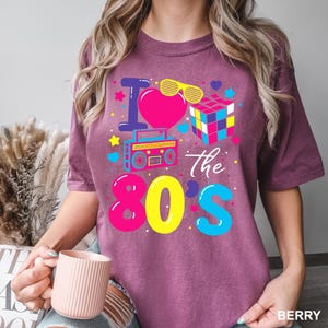 May include: A berry-colored t-shirt featuring a graphic print with the text I Love The 80s. The design includes a pink heart, yellow shutter shades, a colorful puzzle cube, and a boombox, surrounded by small stars, hearts, and dots.