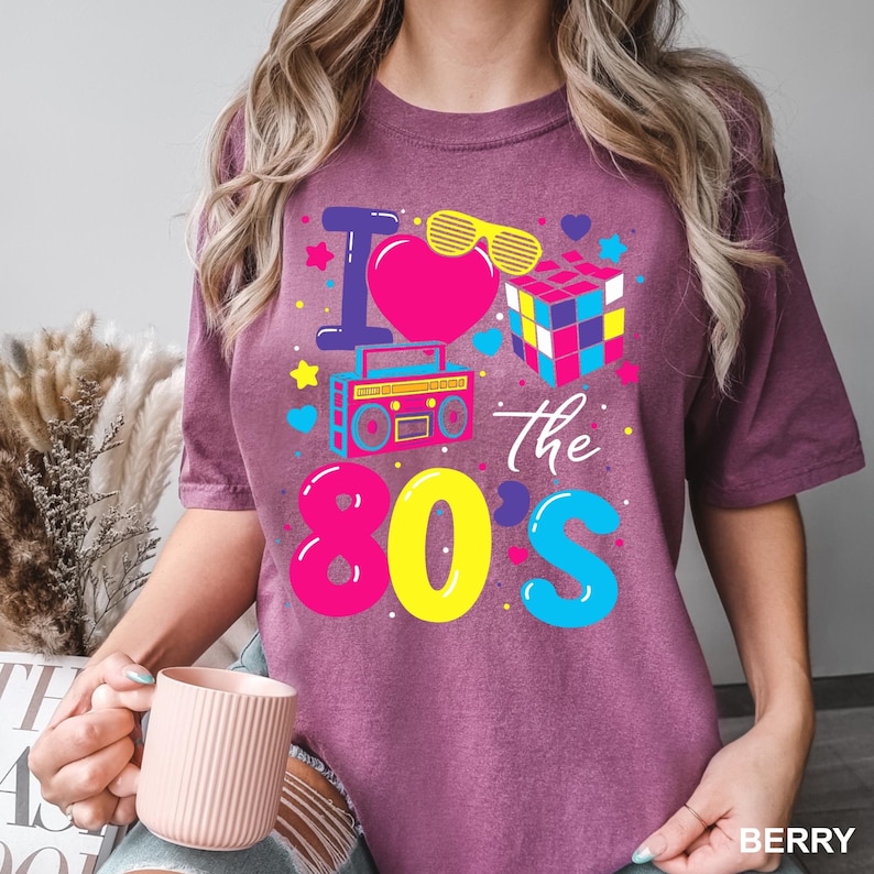 May include: A berry-colored t-shirt featuring a graphic print with the text I Love The 80s. The design includes a pink heart, yellow shutter shades, a colorful puzzle cube, and a boombox, surrounded by small stars, hearts, and dots.