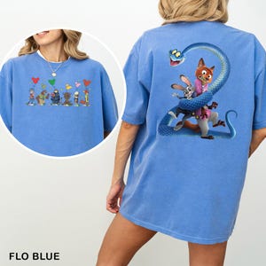 Op de afbeelding: Flo blauw t-shirt met een cartoon illustratie op de voorkant van personages die ballonnen vasthouden. De achterkant heeft een afbeelding van een vos, konijn en slang. De tekst "FLO BLUE" staat onderaan.