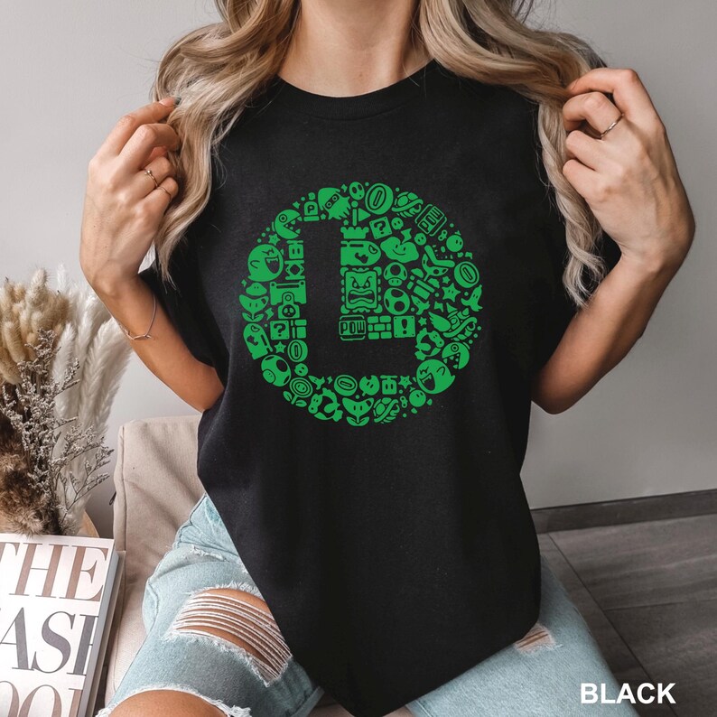 May include: Black t-shirt featuring a green graphic of the letter "L" surrounded by various video game icons. The design includes elements like coins, question marks, and characters. The word "BLACK" is printed at the bottom.